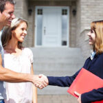 a-young-couple-standing-and-smiling-as-they-shake-hands-with-a-blonde-female-real-estate-on-the-right
