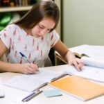 a-young-woman-with-long-dark-hair-is-leaning-over-a-desk-focused-on-her-work-she-is-writing-or-drawing-on-a-large-piece-of-paper-with-blue-pen