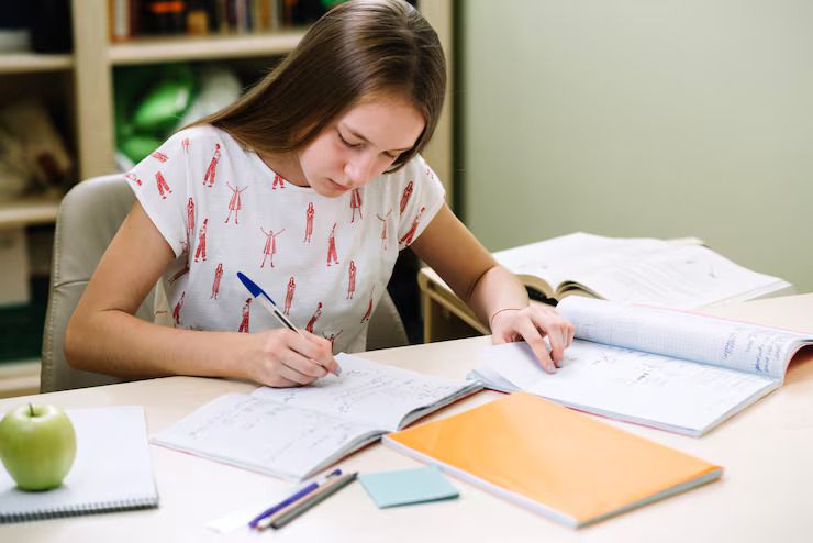a-young-woman-with-long-dark-hair-is-leaning-over-a-desk-focused-on-her-work-she-is-writing-or-drawing-on-a-large-piece-of-paper-with-blue-pen