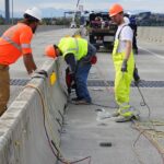 three-construction-workers-wearing-safety-gear-on-a-road-construction-site-and-installing-aligning-or-security-heavy-concrete-barriers-on-a-roadway