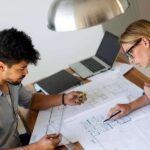 two-people-looking-over-home-layout-plans-documents-at-a-wooden-table