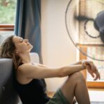 woman-sitting-on-the-floor-in-front-of-a-blue-couch-leaning-back-with-her-head-tilted-upward-and-tall-light-blue-pedestal-fan-aimed-toward-her-cool-down