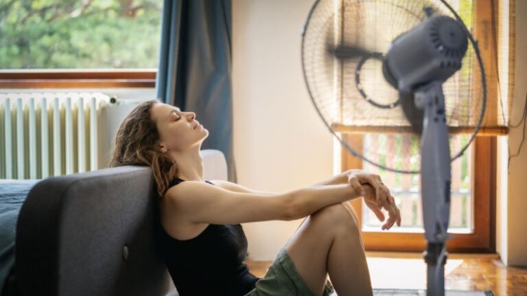 woman-sitting-on-the-floor-in-front-of-a-blue-couch-leaning-back-with-her-head-tilted-upward-and-tall-light-blue-pedestal-fan-aimed-toward-her-cool-down