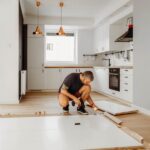 a-man-is-crouching-down-on-the-floor-he-appears-focused-and-is-working-with-his-hands-to-measure-or-lay-down-pieces-of-wood