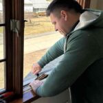 a-man-is-working-on-the-exterior-frame-of-a-dark-wooden-window-he-is-leaning-forward-over-the-window-sill