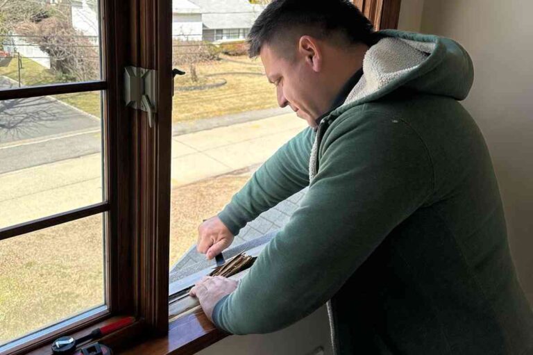 a-man-is-working-on-the-exterior-frame-of-a-dark-wooden-window-he-is-leaning-forward-over-the-window-sill