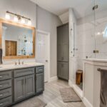 a-modern-bathroom-interior-features-gray-cabinets-and-drawers-a-large-framed-mirror-and-bright-vanity-lightning-above-the-sink