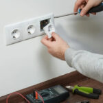 a-person-is-using-a-screwdriver-to-fix-or-install-a-ehite-triple-electrical-outlet-onto-a-wall