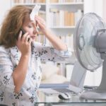 a-woman-is-holding-a-wet-wipe-or-cool-cloth-against-her-forehead-and-sitting-directly-in-front-of-running-fan