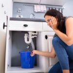 a-woman-sits-on-the-floor-of-kitchen-or-bathroom-she-appears-stressed-holding-her-head-and-looking-at-plumbing-problem