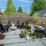 a-circular-stone-paver-patio-holds-three-daek-chairs-surrounded-a-central-light-colored-gas-fire-bowl-in-a-backyard