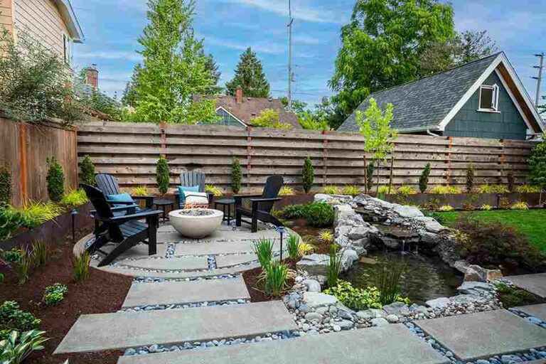 How to Create a Low-Maintenance Backyard That Still Looks High-End a-circular-stone-paver-patio-holds-three-daek-chairs-surrounded-a-central-light-colored-gas-fire-bowl-in-a-backyard