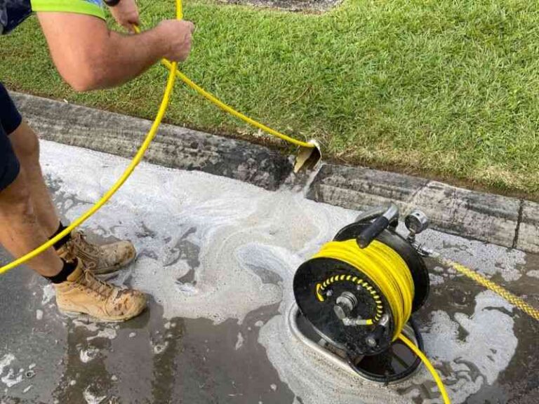 Your Stormwater Drain Looks Clear From Above – Why It’s Still Blocked Underneath a-plumber-is-feeding-a-high-pressure-line-into-an-outdoor-drain-opening-situated-along-the-edge-of-a-green-lawn-and-concrete-surface