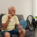 a-elderly-man-sitting-on-a-sofa-during-hot-weather-he-is-wiping-sweat-from-his-face-with-small-white-towel-holding-a-glass-of-water-and-electric-desk-fan-blowing-air-towards-him