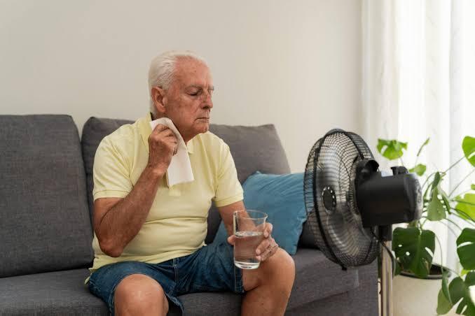 How to Protect Immunocompromised, Elderly and Young Family Members When Heat Waves Strike a-elderly-man-sitting-on-a-sofa-during-hot-weather-he-is-wiping-sweat-from-his-face-with-small-white-towel-holding-a-glass-of-water-and-electric-desk-fan-blowing-air-towards-him