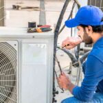 a-male-HVAC-technician-is-kneeling-or-bending-down-to-perform-maintenance-or-a-repair-on-a-large-exterior-air-conditioner-condenser-unit