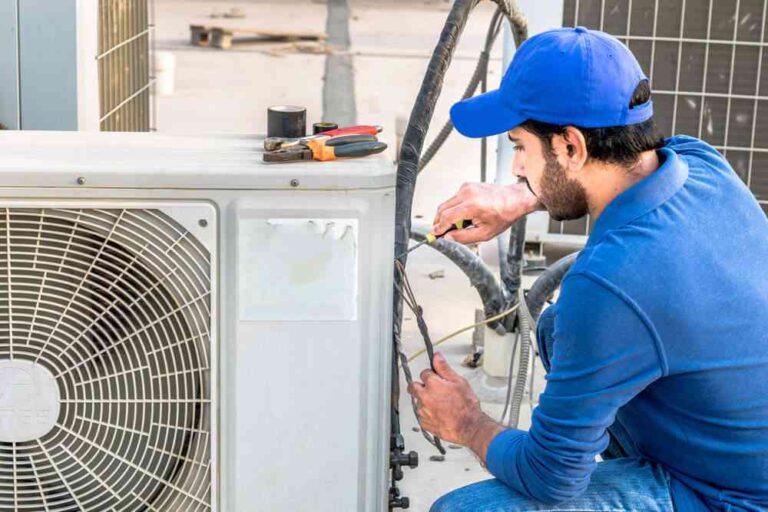 Repair or Replace? 7 Signs It’s Time for a New HVAC System a-male-HVAC-technician-is-kneeling-or-bending-down-to-perform-maintenance-or-a-repair-on-a-large-exterior-air-conditioner-condenser-unit